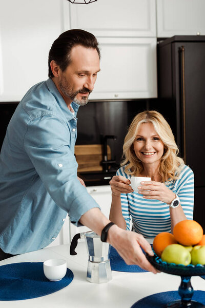 Selective focus of woman with coffee cup smiling at camera near man holding vase with fruits on kitchen table 