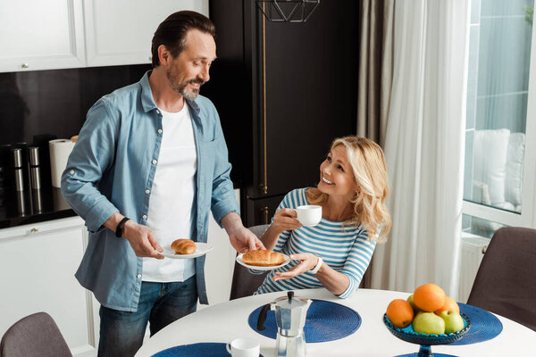 Smiling woman holding cup and looking at husband with croissants in kitchen 