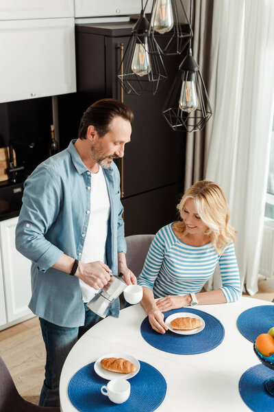 Handsome man pouring coffee in cup near smiling wife at kitchen table 