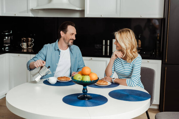 Selective focus of man smiling at wife while pouring coffee near croissants on kitchen table 