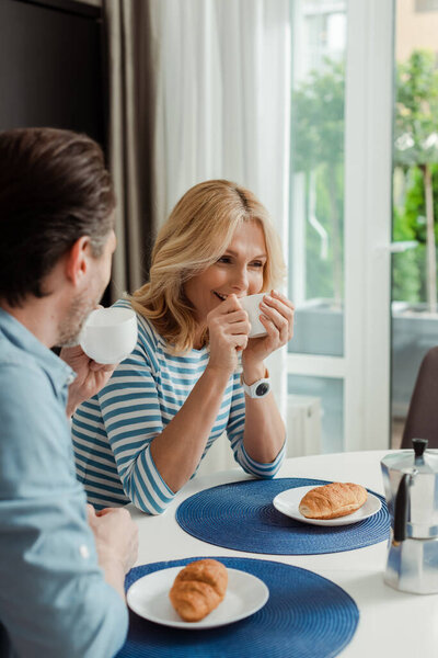 Selective focus of smiling woman drinking coffee near man during breakfast in kitchen 