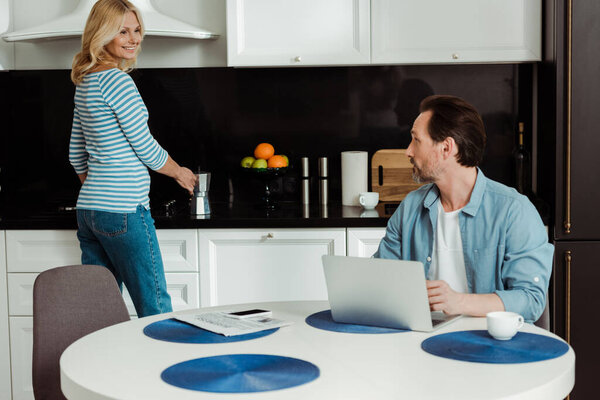 Smiling woman holding geyser coffee maker and smiling at husband using laptop in kitchen 