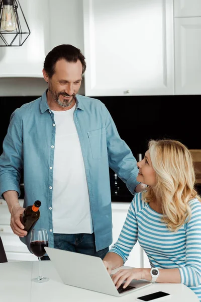 Smiling Man Pouring Wine Wife Using Laptop Kitchen Table — Stock Photo, Image