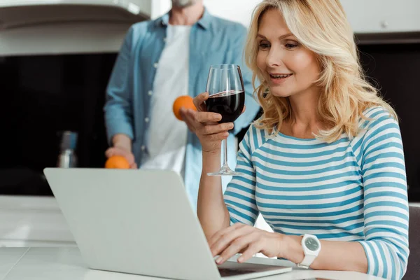 ワインのグラスを保持し キッチンでオレンジを保持夫の近くのノートパソコンを使用して笑顔の女性の選択的な焦点 — ストック写真