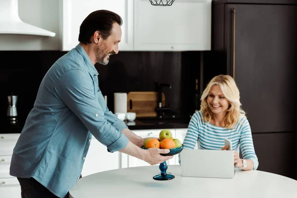 Smiling Man Holding Vase Fruits Wife Using Smartphone Laptop Kitchen — Stock Photo, Image