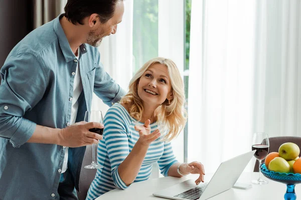 Mujer Sonriente Usando Portátil Señalando Con Dedo Marido Sosteniendo Vaso — Foto de Stock