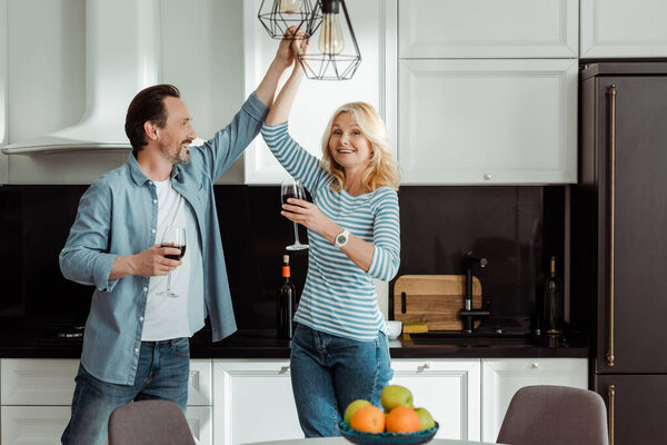 Selective focus of smiling mature couple dancing with glasses of wine in kitchen 