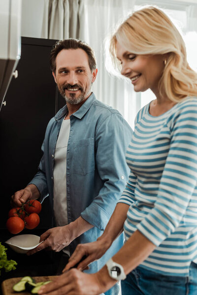 Selective focus of smiling man looking at wife during cooking salad in kitchen 