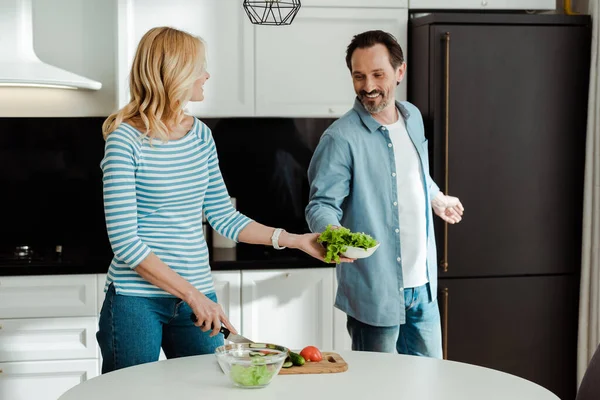台所で野菜を切る妻にレタスを与える笑顔の男 — ストック写真