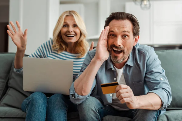 Shocked Mature Man Holding Credit Card Cheerful Wife Laptop Couch — Stock Photo, Image