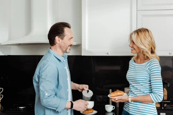 Sorrindo Homem Derramando Café Perto Bela Esposa Segurando Croissant Cozinha — Fotografia de Stock