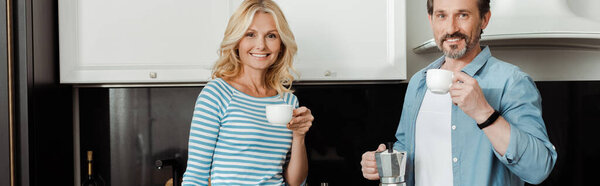 Panoramic crop of smiling mature couple drinking coffee in kitchen 