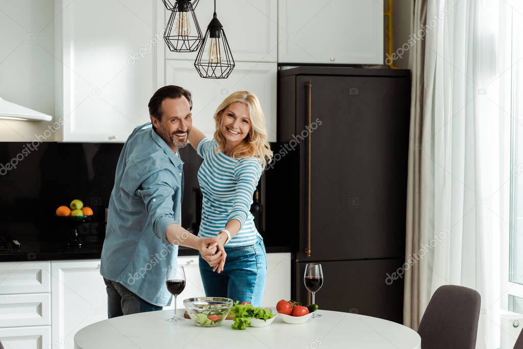 Cheerful mature couple dancing near wine glasses and fresh salad in kitchen 