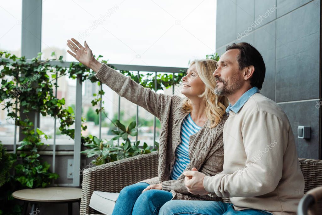 Smiling woman pointing with hand near handsome husband on wicker sofa on terrace