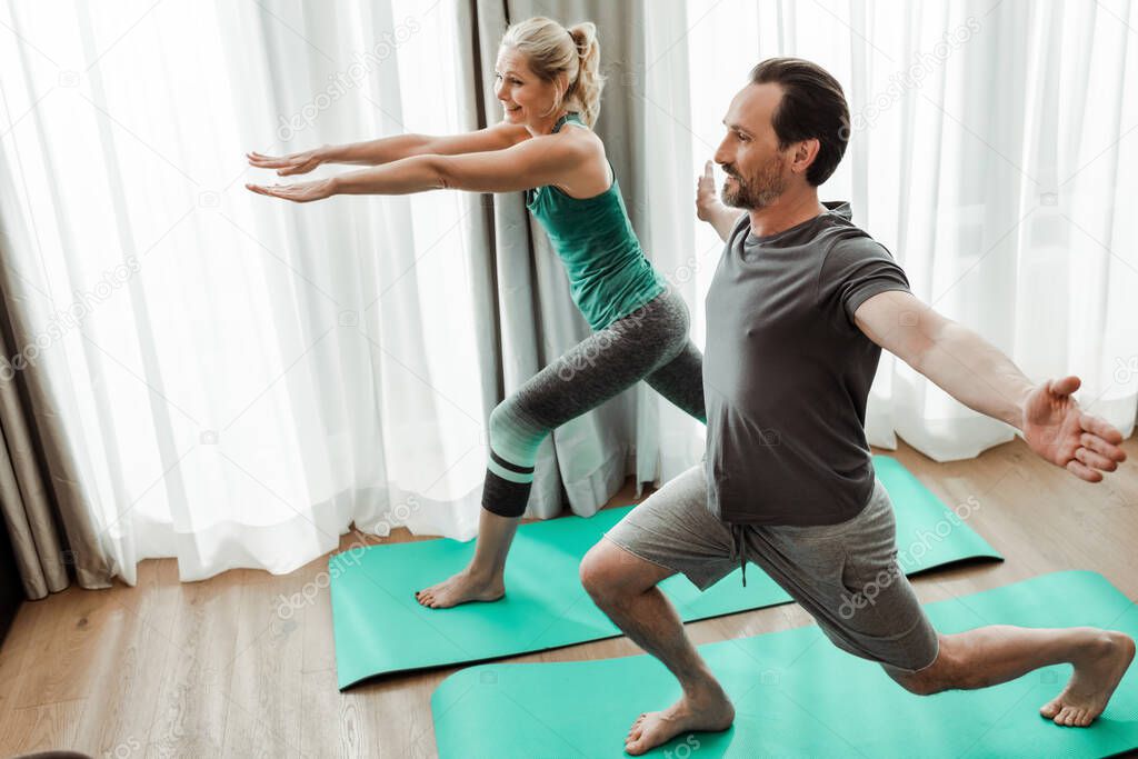 Smiling mature man training on fitness mat near cheerful wife at home 