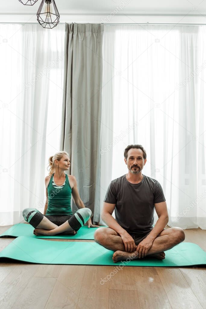 Mature man looking at camera while sitting on fitness mat near wife 