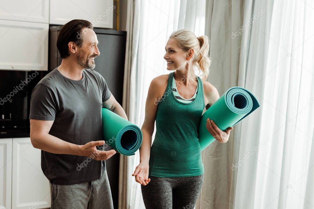 Smiling mature man pointing with hand on fitness mat near beautiful wife at home 