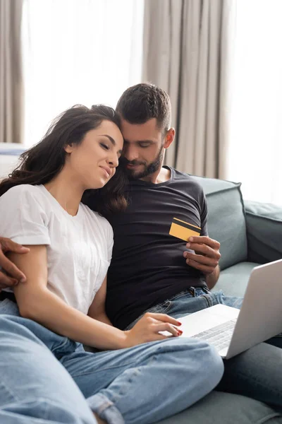 Bonito Homem Segurando Cartão Crédito Abraçando Feliz Namorada Perto Laptop — Fotografia de Stock