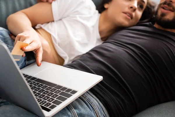 ベッドに寝そべった彼氏とソファに横になりノートパソコンの近くでクレジットカードを持っている女性のクロップビュー — ストック写真