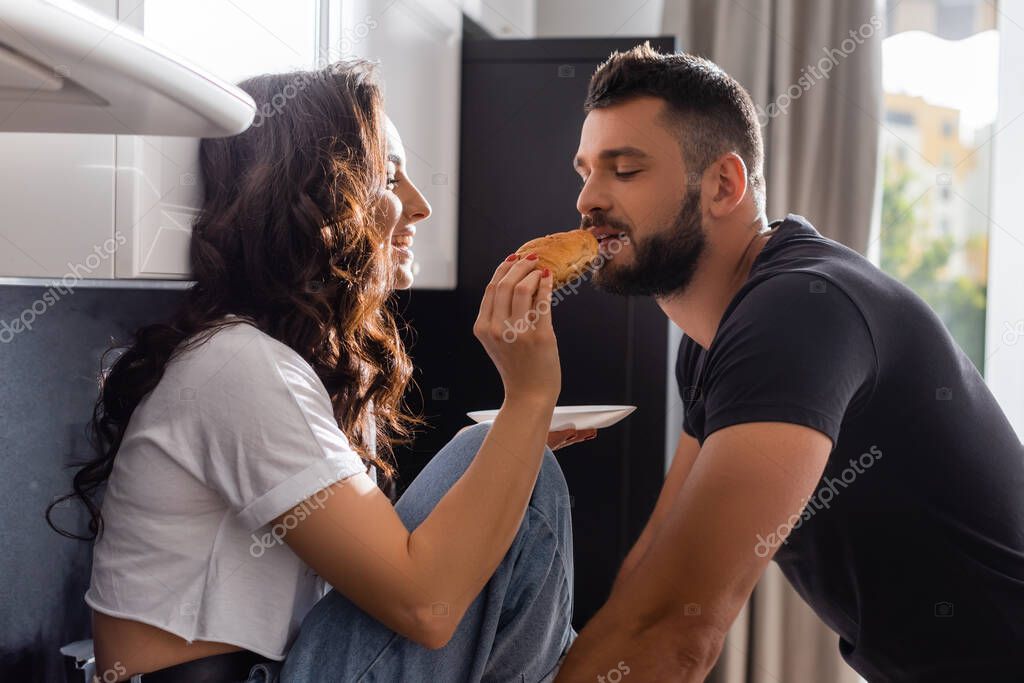 Selective focus of happy girl feeding handsome boyfriend with croissant
