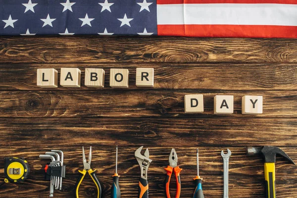 top view of metallic tools and american flag near cubes with labor day lettering on wooden surface 