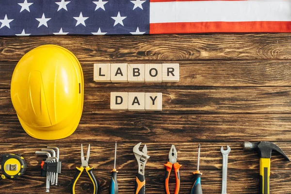 top view of metallic tools, safety helmet, american flag near cubes with labor day lettering on wooden surface