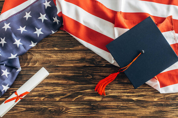 top view of graduation cap and diploma near american flag with stars and stripes on wooden surface 