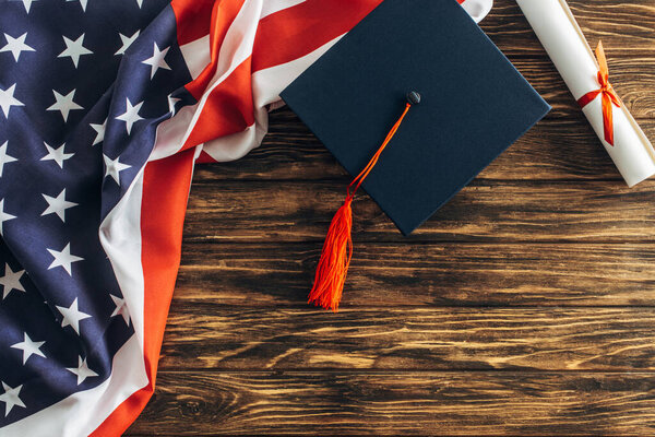 top view of diploma and graduation cap near american flag with stars and stripes on wooden surface 