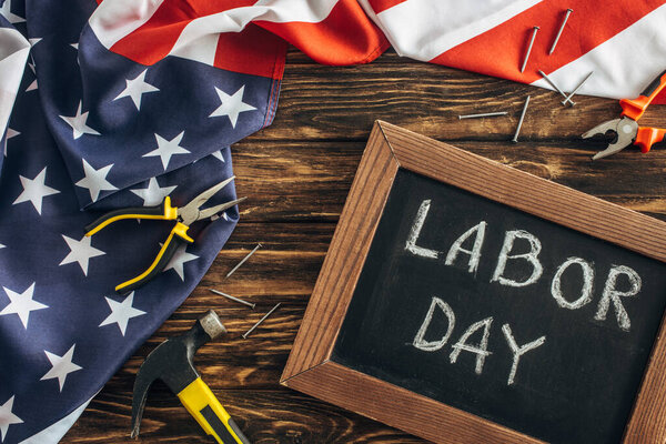 top view of american flag, metallic nails and instruments near chalkboard with labor day lettering on wooden surface 