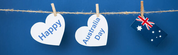 panoramic shot of rope, clothespins and heart-shaped papers with happy near australia day lettering and flag on blue