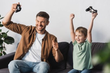 KYIV, UKRAINE - JUNE 9, 2020: excited father and son showing winner gestures while holding joysticks clipart
