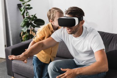 VR kulaklıklı heyecanlı bir adam uzanmış elleriyle oturuyor ve sevimli oğlu onunla konuşuyor.
