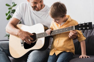 Mutlu bir baba, gülümseyen oğluna akustik gitar çalmayı öğretiyor.