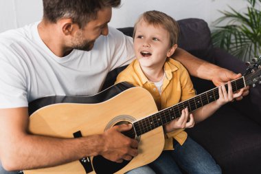 Gülümseyen baba heyecanlı oğula evde akustik gitar çalmayı öğretiyor.