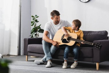 Baba oğul birbirlerine bakarken çocuk akustik gitar çalmayı öğreniyor.