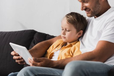 Sevimli oğluyla gülümseyen baba kanepeye otururken dijital tablet kullanıyor.