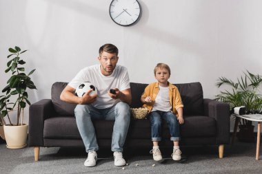 Endişeli adam elinde futbol topu ve uzaktan kumandayla oğlunun yanında patlamış mısır yiyor.