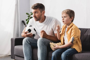 Endişeli baba ve oğul ellerinde futbol topu ve patlamış mısır tutarken televizyon izliyorlar.