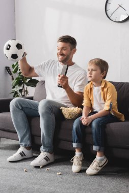 Neşeli adam elinde uzaktan kumandayla futbol topu tutuyor.