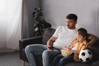 Sıkılmış baba ve oğul futbol topunun yanında patlamış mısırla televizyon izliyorlar.