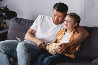 Endişeli baba ve gülümseyen oğul televizyon izliyor ve evdeki kanepede patlamış mısır yiyorlar.