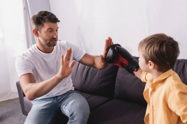 Yakışıklı adam kanepede oturuyor ve oğluyla boks eldiveni giyerek kavga ediyor.
