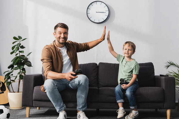 KYIV, UKRAINE - JUNE 9, 2020: happy father and son giving high and smiling at camera five while holding joysticks 