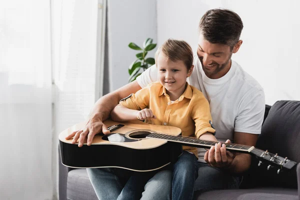 Smiling Boy Sitting Fathers Knees Learning How Play Acoustic Guitar — Stock Photo, Image