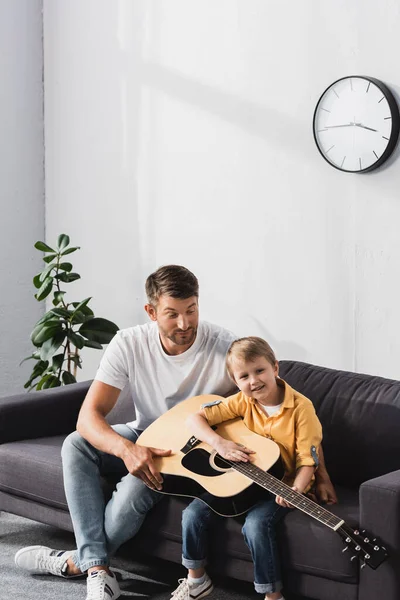 Jóképű Apa Tanít Fia Játszani Akusztikus Gitár Kanapén Otthon — Stock Fotó