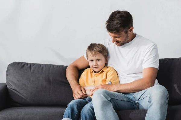 Šťastný Otec Objímání Rozkošný Syn Zatímco Sedí Pohovce Doma — Stock fotografie