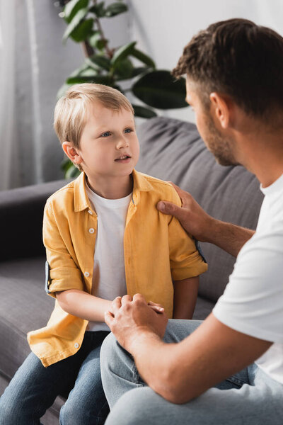 caring father touching shoulder and holding hand of adorable son while talking to him at home