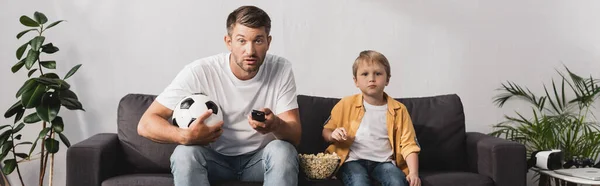 息子の近くでサッカーボールとテレビのリモコンを持っている心配男の水平画像ポップコーンを食べて — ストック写真