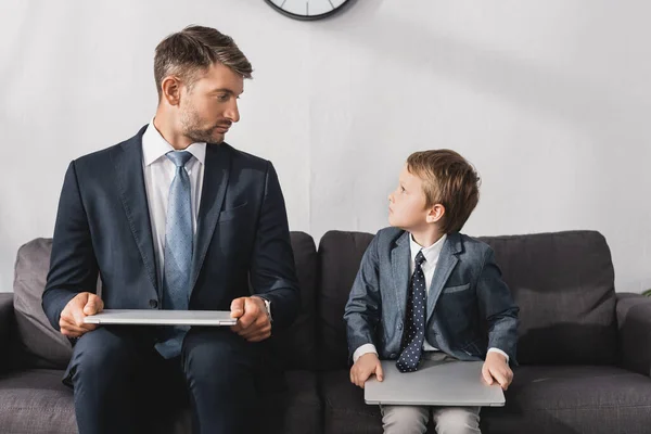 认真的商人和他的儿子穿着正式的衣服 坐在家里沙发上的笔记本电脑上 — 图库照片