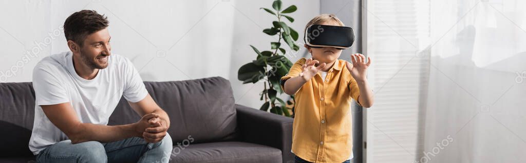 horizontal image of boy in vr headset standing with outstretched hands near father sitting on sofa